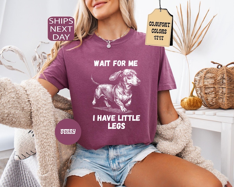 May include: Berry-colored t-shirt with a dachshund graphic and the text "WAIT FOR ME I HAVE LITTLE LEGS." The shirt is a Comfort Colors 1717 style. The model is wearing denim shorts and a cream-colored cardigan.