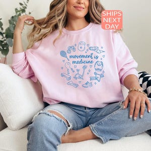 May include: A light pink sweatshirt with the text "movement is medicine" in a circle of blue exercise-related graphics. The sweatshirt has long sleeves and a crew neck. The image also includes the text "SHIPS NEXT DAY".