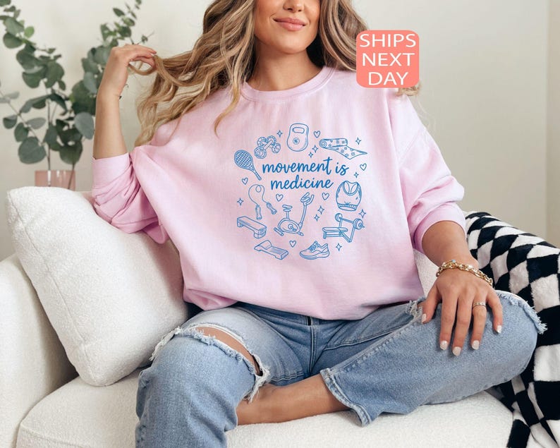 May include: A light pink sweatshirt with the text "movement is medicine" in a circle of blue exercise-related graphics. The sweatshirt has long sleeves and a crew neck. The image also includes the text "SHIPS NEXT DAY".