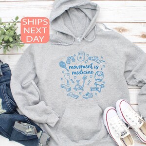 May include: A light gray hoodie with a blue graphic of fitness equipment and the words "movement is medicine." The hoodie is paired with blue jeans and white sneakers. A small plant is visible in the background.