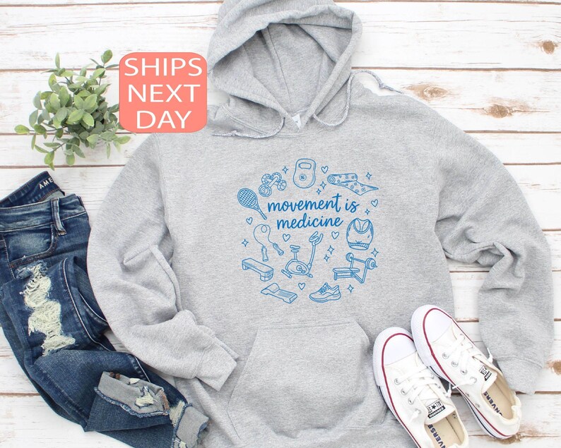 May include: A light gray hoodie with a blue graphic of fitness equipment and the words "movement is medicine." The hoodie is paired with blue jeans and white sneakers. A small plant is visible in the background.