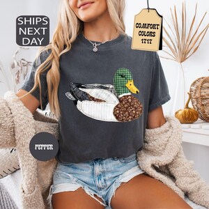 May include: Dark gray t-shirt featuring a duck design with a green plaid head, brown floral body, and white striped wings. The shirt has the text "COMFORT COLORS 1717" on a tag. The shirt also has the text "PEPPER" on a circle.