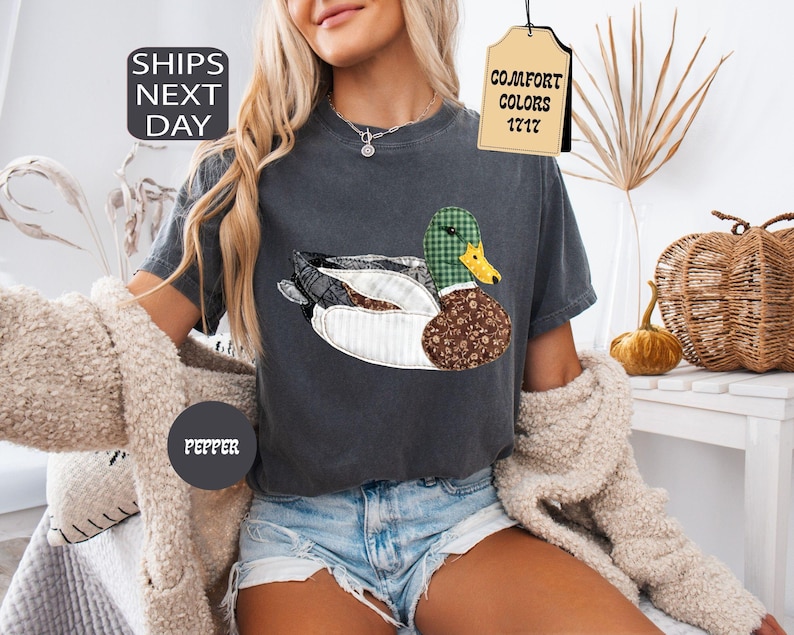 May include: Dark gray t-shirt featuring a duck design with a green plaid head, brown floral body, and white striped wings. The shirt has the text "COMFORT COLORS 1717" on a tag. The shirt also has the text "PEPPER" on a circle.