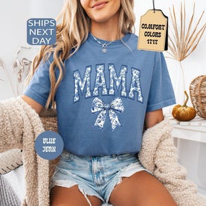May include: A blue short-sleeved t-shirt featuring the word MAMA in a collegiate font with a white and blue floral pattern. Below the text is a large bow graphic with the same floral design. The shirt has a crew neckline and a textured fabric finish.