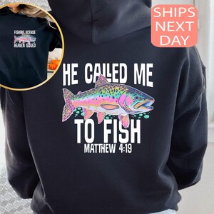 Peut inclure: Sweat &agrave; capuche noir avec un motif de poisson color&eacute; et le texte "HE CALLED ME TO FISH MATTHEW 4:19." Une petite image en haut &agrave; gauche montre une licence de p&ecirc;che avec le texte "FISHING LICENSE HEAVEN ISSUED."