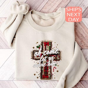 Peut inclure: Sweat-shirt crème avec un motif de croix et les mots "oh come let us adore him". La croix est ornée de carreaux rouges, de houx et d'étoiles dorées. Le sweat-shirt a un col rond et des manches longues.