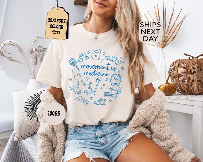 K&ouml;nnte beinhalten: Elfenbeinfarbenes T-Shirt mit einer blauen Grafik von Fitnessger&auml;ten und dem Schriftzug "movement is medicine". Das Shirt wird mit Jeansshorts getragen. Das Bild enth&auml;lt auch eine beige Strickjacke, ein dekoratives Kissen und einen Weidenkorb.