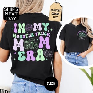 May include: A black t-shirt featuring a graphic on the back with the text IN MY MONSTER TRUCK MOM ERA in wavy light purple and mint green letters, accompanied by monster truck illustrations and stars. The front chest has a small monster truck graphic.