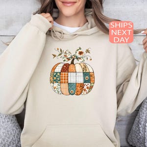 May include: Beige hoodie featuring a patchwork pumpkin design. The pumpkin is composed of various fabric patterns in orange, green, and white, topped with a bow and floral accents. The text "SHIPS NEXT DAY" is displayed on a pink square.