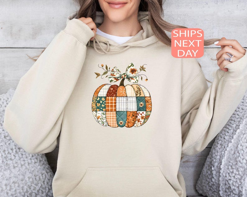 May include: Beige hoodie featuring a patchwork pumpkin design. The pumpkin is composed of various fabric patterns in orange, green, and white, topped with a bow and floral accents. The text "SHIPS NEXT DAY" is displayed on a pink square.