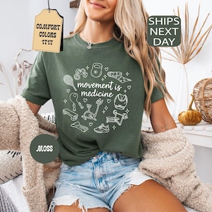 K&ouml;nnte beinhalten: Ein moosgr&uuml;nes T-Shirt mit dem Schriftzug "movement is medicine" in einem wei&szlig;en kreisf&ouml;rmigen Design mit Fitnessger&auml;ten. Das Shirt hat einen Rundhalsausschnitt und kurze &Auml;rmel. Das Shirt wird mit Jeansshorts kombiniert.