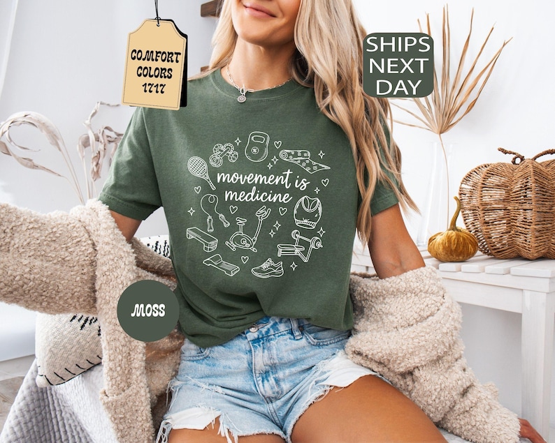 K&ouml;nnte beinhalten: Ein moosgr&uuml;nes T-Shirt mit dem Schriftzug "movement is medicine" in einem wei&szlig;en kreisf&ouml;rmigen Design mit Fitnessger&auml;ten. Das Shirt hat einen Rundhalsausschnitt und kurze &Auml;rmel. Das Shirt wird mit Jeansshorts kombiniert.