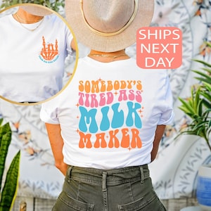 May include: White t-shirt with the text "SOMEBODY'S TIRED ASS MILK MAKER" in retro orange, pink, blue, and orange lettering. The front of the shirt has a skeleton hand graphic with the text "RAISE HELL BABE".