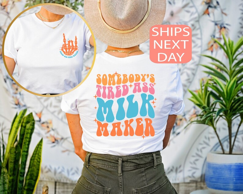 May include: White t-shirt with the text "SOMEBODY'S TIRED ASS MILK MAKER" in retro orange, pink, blue, and orange lettering. The front of the shirt has a skeleton hand graphic with the text "RAISE HELL BABE".