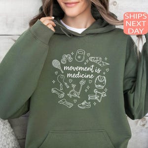 May include: An olive green hoodie with a white graphic of exercise equipment, including a dumbbell, yoga mat, and jump rope. The text "movement is medicine" is centered within the graphic. The hoodie has a front pocket and a drawstring hood.