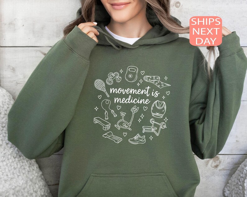 May include: An olive green hoodie with a white graphic of exercise equipment, including a dumbbell, yoga mat, and jump rope. The text "movement is medicine" is centered within the graphic. The hoodie has a front pocket and a drawstring hood.