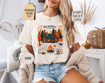 Op de afbeelding: Een ivoorkleurig t-shirt met een grafische print van een blokhut in een bos met herfstbomen, een kampvuur, open boeken, een mok en een pompoen. De tekst luidt READING RETREAT, cozy cabin, a relaxing bookish escape, for the cozy readers.