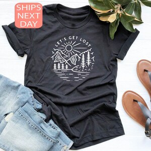 May include: Black t-shirt with a white graphic of a mountain scene, sun, and the text "LET'S GET LOST". The shirt is paired with blue denim jeans and brown sandals. The image also includes a sprig of green leaves.