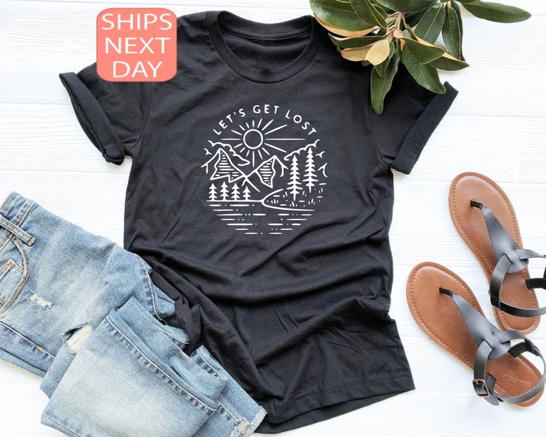 May include: Black t-shirt with a white graphic of a mountain scene, sun, and the text "LET'S GET LOST". The shirt is paired with blue denim jeans and brown sandals. The image also includes a sprig of green leaves.