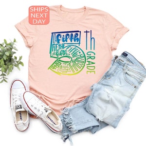 Pu&ograve; includere: T-shirt rosa chiaro con grafica colorata "Fifth Grade" in sfumature di blu, verde e giallo. L'immagine include jeans chiari e scarpe da ginnastica bianche, suggerendo un tema di ritorno a scuola.