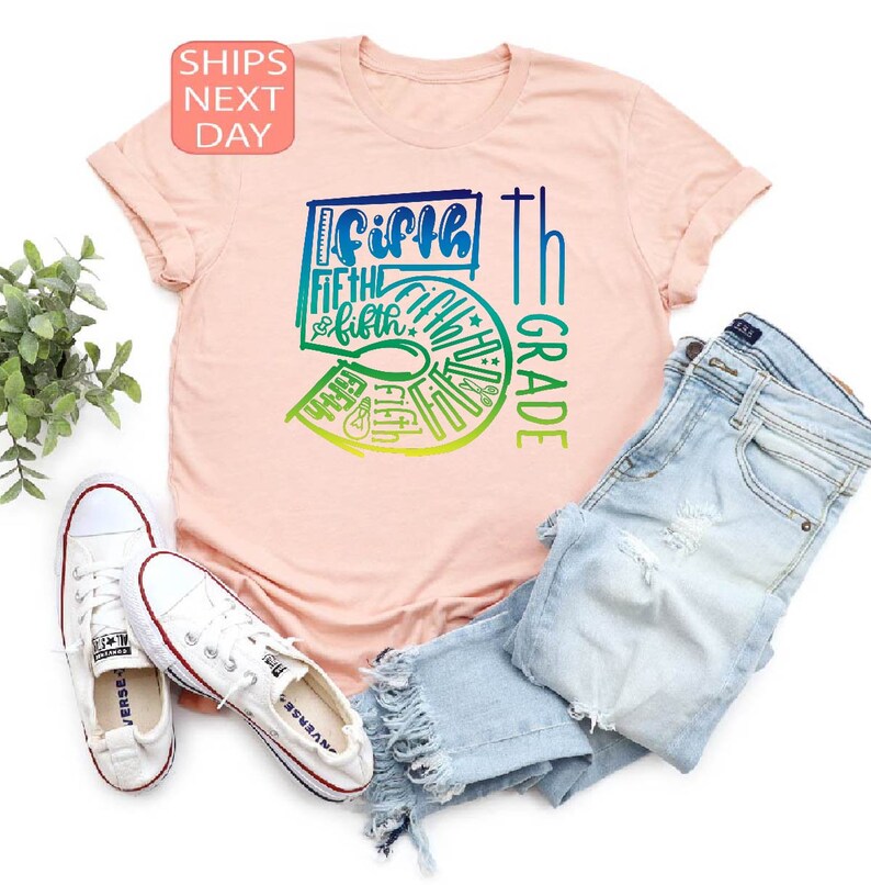 Pu&ograve; includere: T-shirt rosa chiaro con grafica colorata "Fifth Grade" in sfumature di blu, verde e giallo. L'immagine include jeans chiari e scarpe da ginnastica bianche, suggerendo un tema di ritorno a scuola.