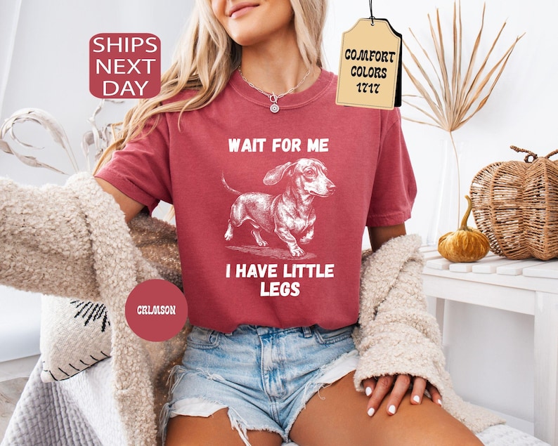 May include: Crimson red t-shirt with a white illustration of a dachshund and the text "WAIT FOR ME I HAVE LITTLE LEGS". The shirt also has the text "COMFORT COLORS 1717". The model is wearing denim shorts.