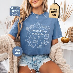 K&ouml;nnte beinhalten: Blaues Jeans-T-Shirt mit dem Schriftzug "movement is medicine" und wei&szlig;en Illustrationen von Trainingsger&auml;ten. Das Shirt hat einen Rundhalsausschnitt und kurze &Auml;rmel. Das Shirt ist Teil der Comfort Colors 1717 Kollektion.