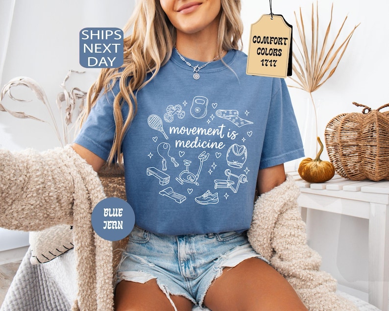 K&ouml;nnte beinhalten: Blaues Jeans-T-Shirt mit dem Schriftzug "movement is medicine" und wei&szlig;en Illustrationen von Trainingsger&auml;ten. Das Shirt hat einen Rundhalsausschnitt und kurze &Auml;rmel. Das Shirt ist Teil der Comfort Colors 1717 Kollektion.