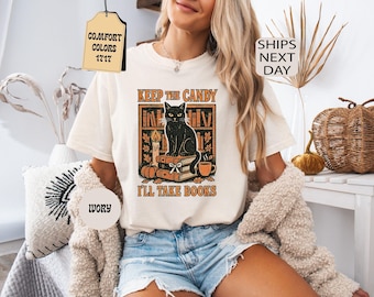 May include: An ivory t-shirt featuring a graphic of a black cat sitting on a stack of books in front of a bookshelf. The design includes a candle, a pumpkin, and a mug, with the text "KEEP THE CANDY I'LL TAKE BOOKS" printed in orange letters.