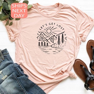 May include: A light pink t-shirt with a round neckline. The shirt features a black graphic of a mountain scene with the text "LET'S GET LOST" in a circular design. The shirt is paired with blue jeans and black sandals.
