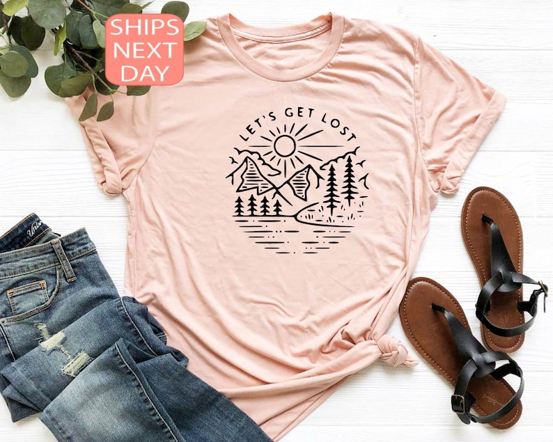 May include: A light pink t-shirt with a round neckline. The shirt features a black graphic of a mountain scene with the text "LET'S GET LOST" in a circular design. The shirt is paired with blue jeans and black sandals.