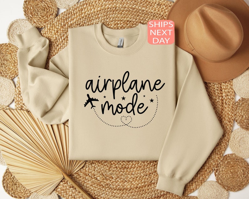 K&ouml;nnte beinhalten: Ein beiger Pullover mit dem Text "airplane mode" in schwarzer Kursivschrift. Neben dem Text befindet sich ein kleines Flugzeugsymbol mit Sternen und einer gepunkteten Linie darum herum.