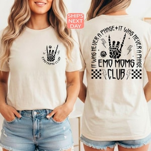 May include: A cream-coloured t-shirt with black graphic designs. The front displays a hand gesture and the text "EMO MOMS CLUB". The back has the text "IT WAS NEVER A PHASE" and a similar hand gesture design. The shirt is short-sleeved.