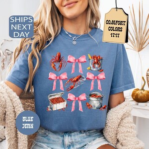 K&ouml;nnte beinhalten: Ein blaues T-Shirt mit einem Motiv aus Krebsen, T&ouml;pfen und rosafarbenen Schleifen. Der Text "Comfort Colors 1717" befindet sich auf einem Etikett, das am Shirt befestigt ist.
