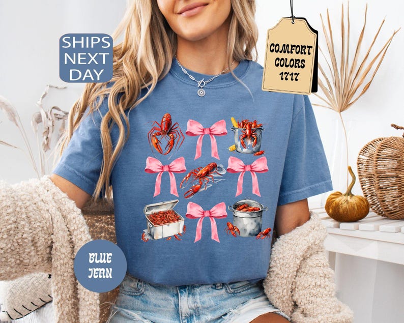 K&ouml;nnte beinhalten: Ein blaues T-Shirt mit einem Motiv aus Krebsen, T&ouml;pfen und rosafarbenen Schleifen. Der Text "Comfort Colors 1717" befindet sich auf einem Etikett, das am Shirt befestigt ist.