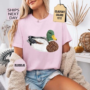 May include: A light pink t-shirt featuring a detailed duck design with a green plaid head, brown patterned body, and white accents. The shirt has the text "Comfort Colors 1717" on a tag. The word "Blossom" is also visible.