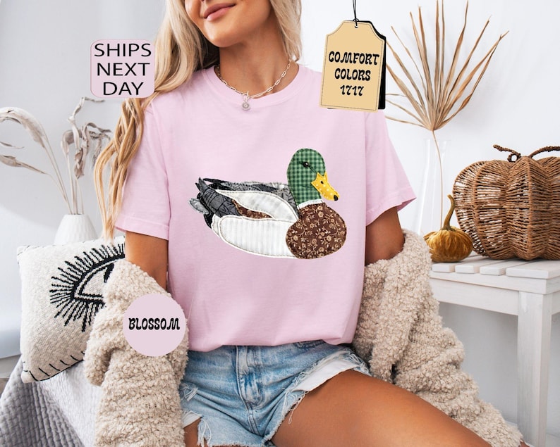 May include: A light pink t-shirt featuring a detailed duck design with a green plaid head, brown patterned body, and white accents. The shirt has the text "Comfort Colors 1717" on a tag. The word "Blossom" is also visible.