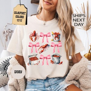 K&ouml;nnte beinhalten: Ein wei&szlig;es T-Shirt mit einem Aufdruck von rosa Schleifen und roten Krebsen in T&ouml;pfen. Ein Etikett, das am Shirt befestigt ist, tr&auml;gt die Aufschrift "COMFORT COLORS 1717".
