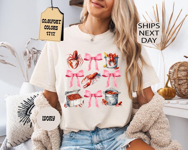 K&ouml;nnte beinhalten: Ein wei&szlig;es T-Shirt mit einem Aufdruck von rosa Schleifen und roten Krebsen in T&ouml;pfen. Ein Etikett, das am Shirt befestigt ist, tr&auml;gt die Aufschrift "COMFORT COLORS 1717".
