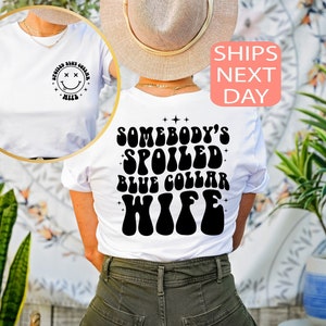 以下が含まれることがあります： 白地に黒のスマイリーフェイス（目はX）がプリントされたTシャツ。後ろにはレトロなフォントで「Somebody's Spoiled Blue Collar Wife」と書いてある。