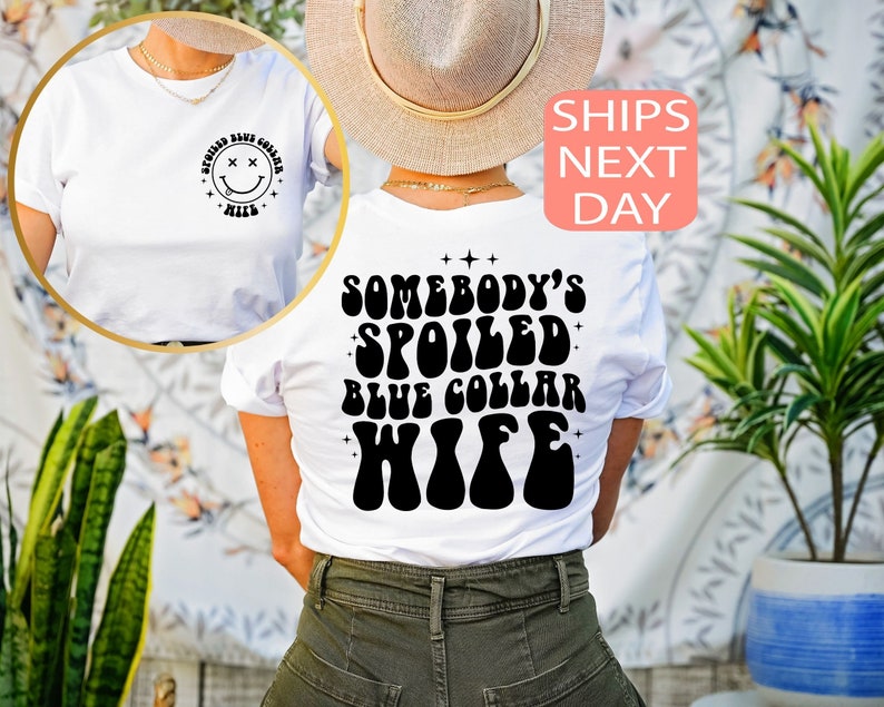 以下が含まれることがあります： 白地に黒のスマイリーフェイス（目はX）がプリントされたTシャツ。後ろにはレトロなフォントで「Somebody's Spoiled Blue Collar Wife」と書いてある。