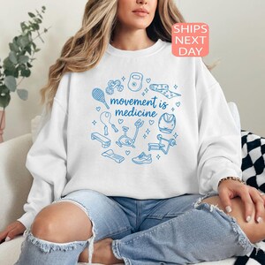 May include: White sweatshirt with a blue circular design featuring fitness equipment and the text "movement is medicine." The sweatshirt is worn with ripped blue jeans. A pink and orange "SHIPS NEXT DAY" sticker is in the top right corner.