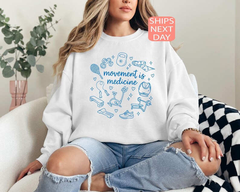 May include: White sweatshirt with a blue circular design featuring fitness equipment and the text "movement is medicine." The sweatshirt is worn with ripped blue jeans. A pink and orange "SHIPS NEXT DAY" sticker is in the top right corner.