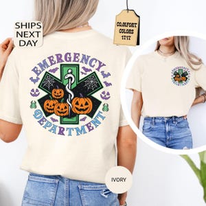 May include: An ivory t-shirt featuring a large graphic on the back and a smaller version on the front left chest. The design displays a green Star of Life symbol with three orange jack-o'-lanterns, spiderwebs, bats, and the text EMERGENCY DEPARTMENT.