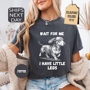 May include: Dark gray t-shirt featuring a dachshund illustration with the text "WAIT FOR ME I HAVE LITTLE LEGS". The shirt has a vintage look and is labeled "Pepper" and "Comfort Colors 1717".