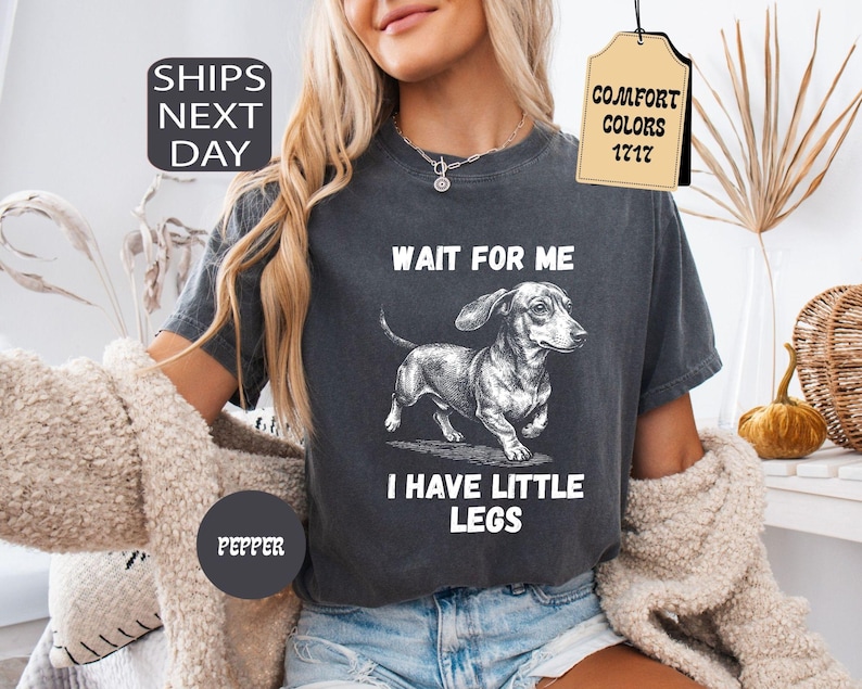 May include: Dark gray t-shirt featuring a dachshund illustration with the text "WAIT FOR ME I HAVE LITTLE LEGS". The shirt has a vintage look and is labeled "Pepper" and "Comfort Colors 1717".