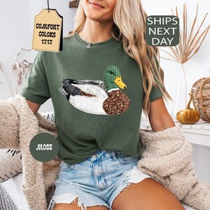 May include: Moss green t-shirt featuring a patchwork duck design with a green plaid head and brown patterned body. The shirt has the text "Comfort Colors 1717" on a tag. The shirt is part of the Comfort Colors collection.