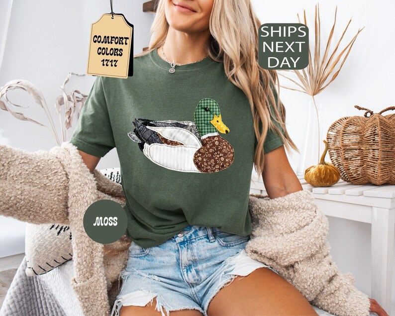 May include: Moss green t-shirt featuring a patchwork duck design with a green plaid head and brown patterned body. The shirt has the text "Comfort Colors 1717" on a tag. The shirt is part of the Comfort Colors collection.