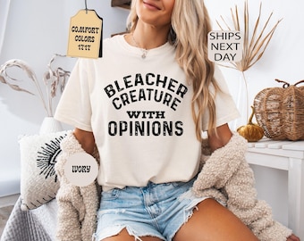Puede incluir: Una camiseta de manga corta color marfil con el texto negro "BLEACHER CREATURE WITH OPINIONS" impreso en una fuente negrita y desgastada en el pecho. La camiseta tiene cuello redondo y un corte relajado, combinada con pantalones cortos de mezclilla azul claro.