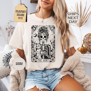 May include: An ivory t-shirt featuring a black graphic print of a skeleton with a high bun hairstyle, holding a cup, accompanied by a cat. The design is framed in a rectangular border with floral accents and the text THE AUNT at the bottom.
